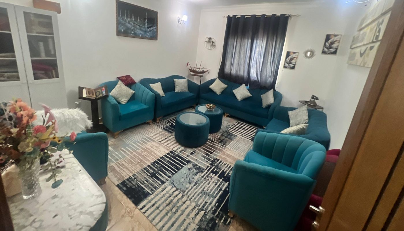 A modern living room with teal sofas, a round ottoman, a patterned rug, wall art, and a large window with dark curtains. A flower vase sits on a marble table in the corner.