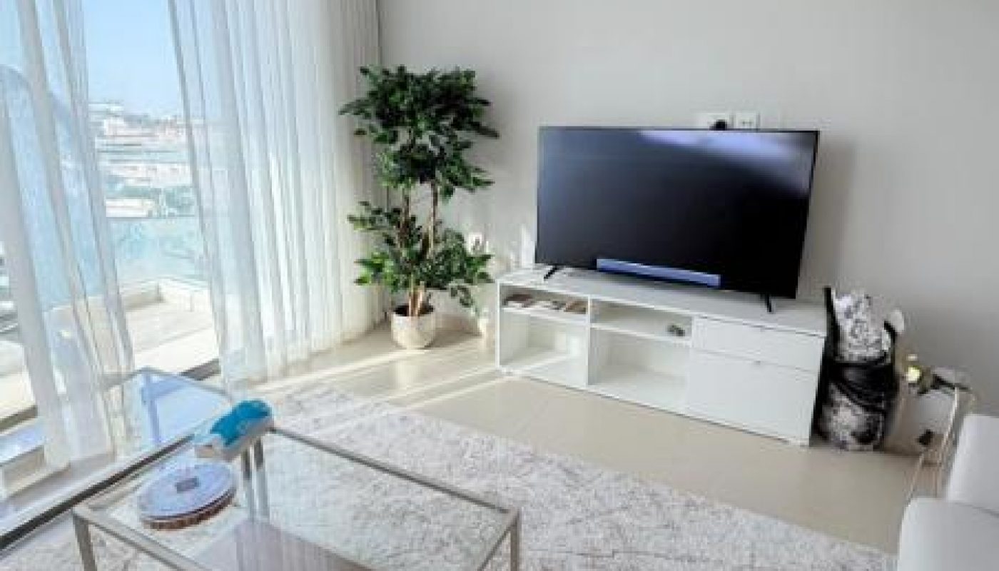 A modern living room with a TV on a white stand, glass coffee table, white rug, large window with sheer curtains, a potted plant, and a view of buildings outside.