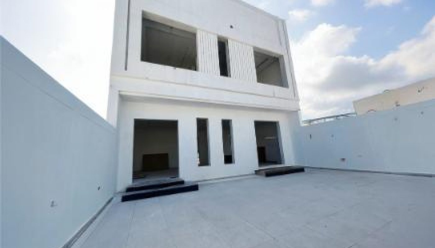 A modern, white two-story building with large windows and a spacious, empty tiled courtyard under a partly cloudy sky.