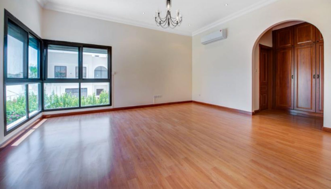 Empty living room with light wood flooring, large corner windows, a chandelier, wall-mounted air conditioner, and built-in wooden closet seen through an arched doorway.