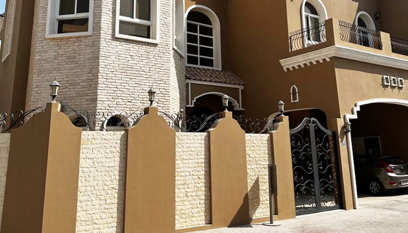 A large, two-story house with tan walls, arched windows, a turret, decorative iron gate, and a car parked in the driveway under a carport.