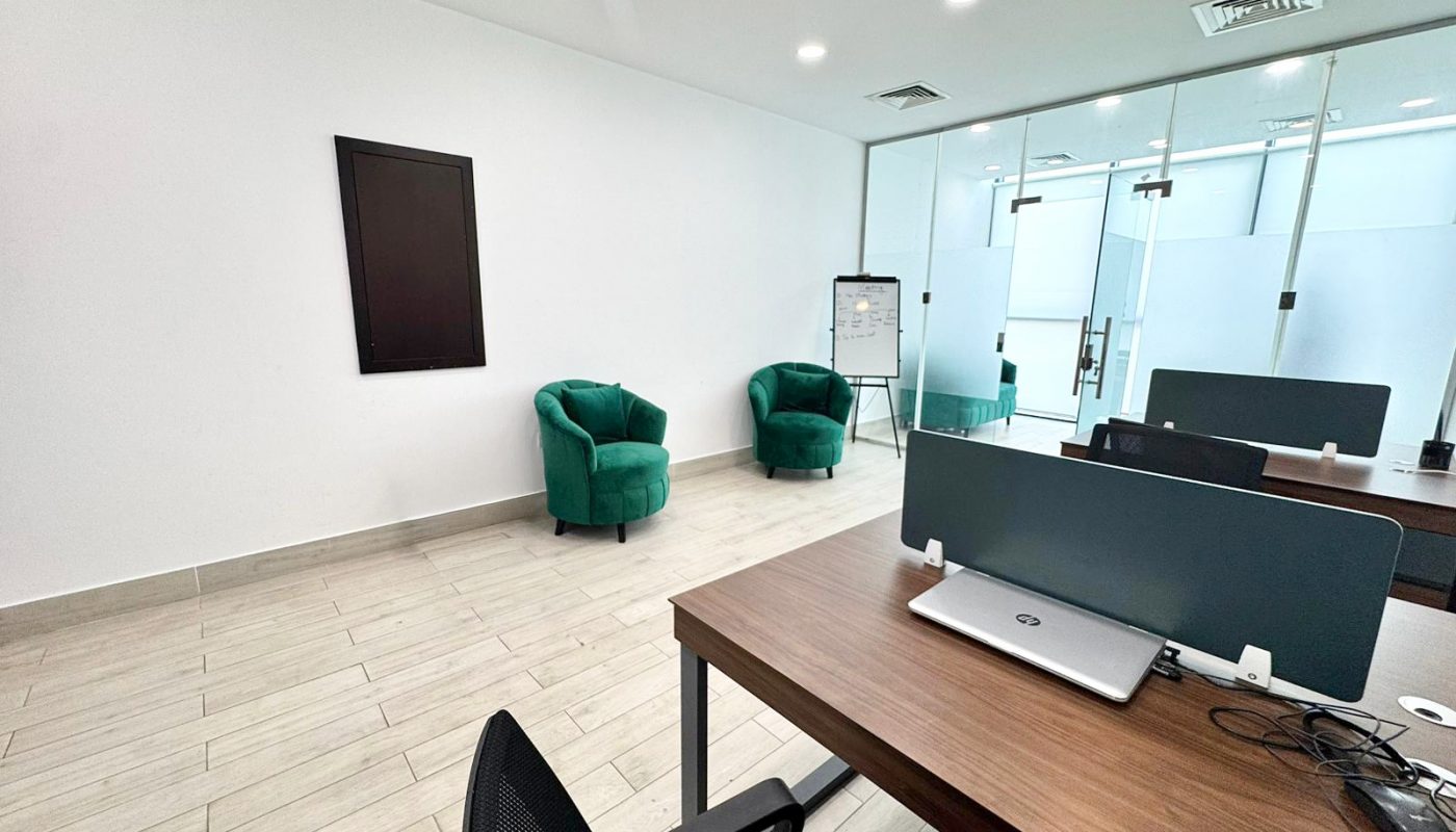 Modern office with two green armchairs against a white wall, desks with laptops in the foreground, and a flipchart near glass doors in the background.