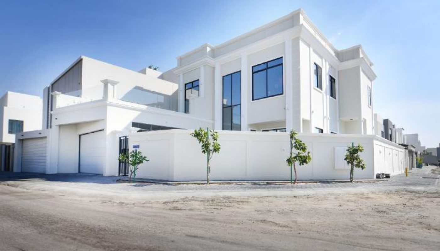 Modern two-story white building with large windows, surrounded by a low wall and a few small trees, set on a paved street under a clear blue sky.