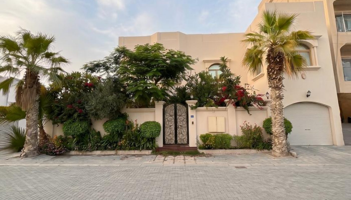 Modern white house with a black gate, surrounded by lush greenery and palm trees, situated next to a paved driveway under a partly cloudy sky.