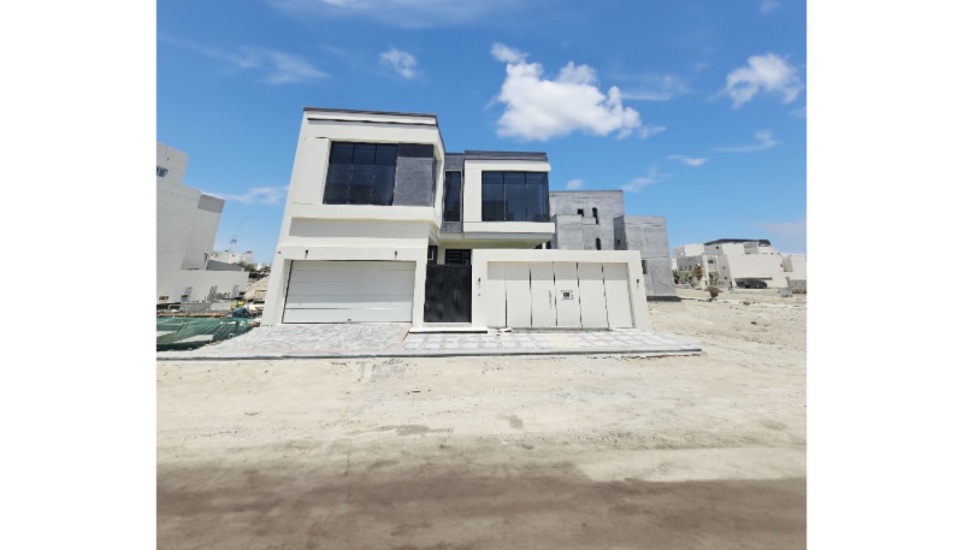 A modern two-story house with large windows and a garage, set on a sandy lot under a blue sky with scattered clouds.