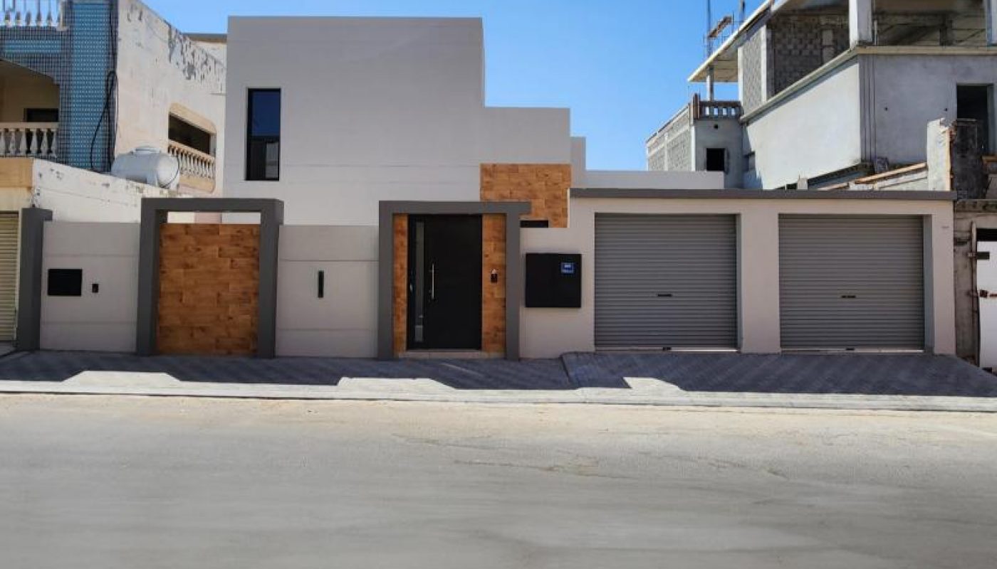 Modern house with a flat roof, white and wood-accented facade, three metal garage doors, and a central entrance, situated on a quiet street under a clear blue sky.