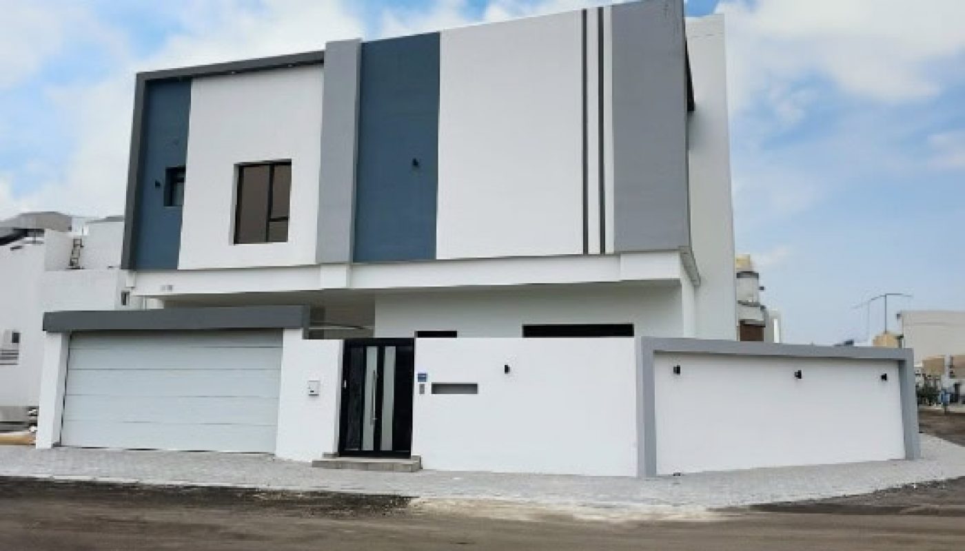 Modern two-story house with a flat roof, white and blue-gray accents, large windows, and a garage. The property is enclosed by a white wall with a black gate. Sky is partly cloudy.
