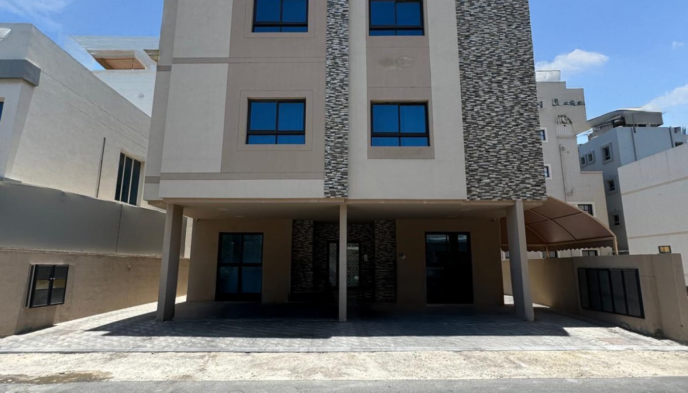 A three-story modern building with a stone accent facade, flat roof, and four large windows on each floor, situated on a paved lot under a blue sky with scattered clouds.