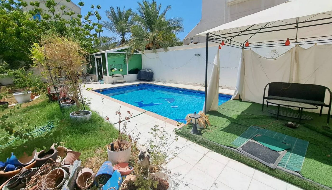 A residential backyard with a swimming pool, a shaded seating area, assorted plants, and scattered gardening supplies looks meticulously designed as if it were an Auto Draft. A large white house stands gracefully in the background.