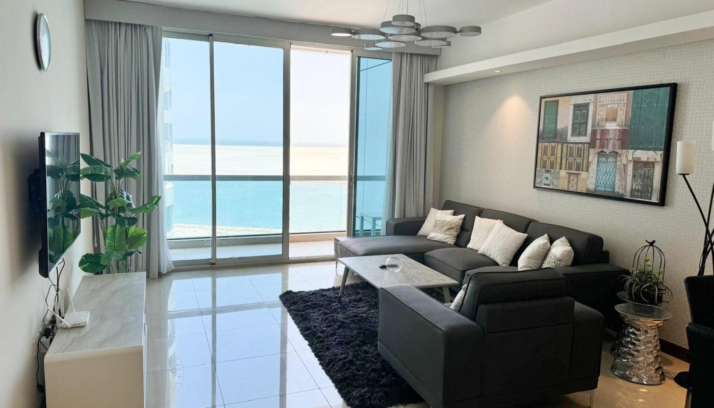 Modern living room with a black sofa set, wall art, and large window offering an ocean view. Marble flooring and a TV mounted on the wall are also visible.