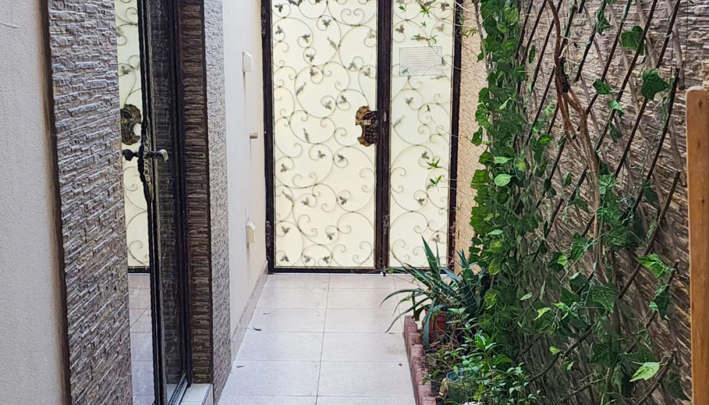 A narrow outdoor corridor with a tiled floor, a small garden bed on the right, and a decorative metal gate at the end.