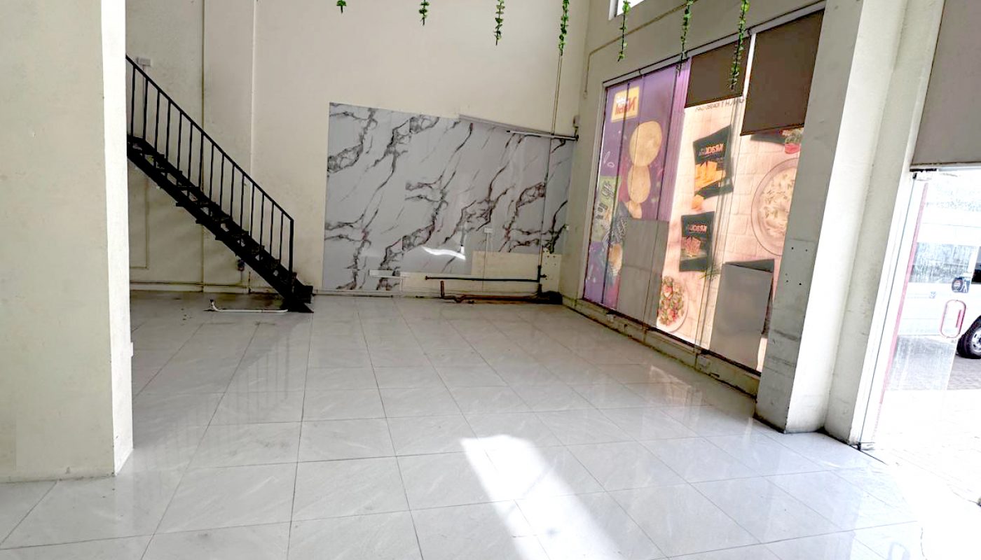Empty commercial space with white marble tile flooring, high ceiling, staircase to upper level, hanging green vines, and large windows with partially covered signage.