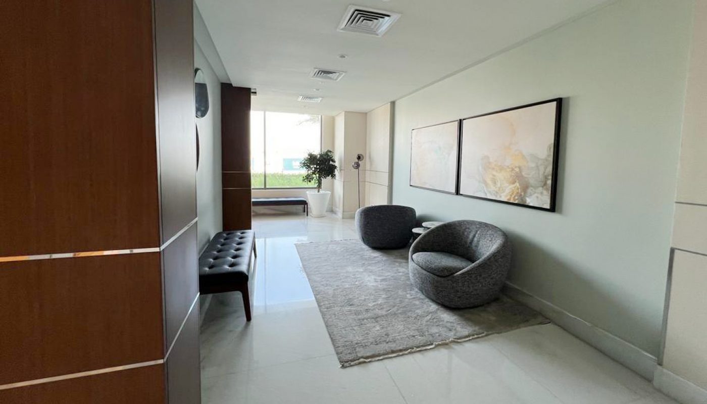A hallway with white floors, two round gray chairs on a rug, a black bench, large wall art, and a potted plant near a window at the end.