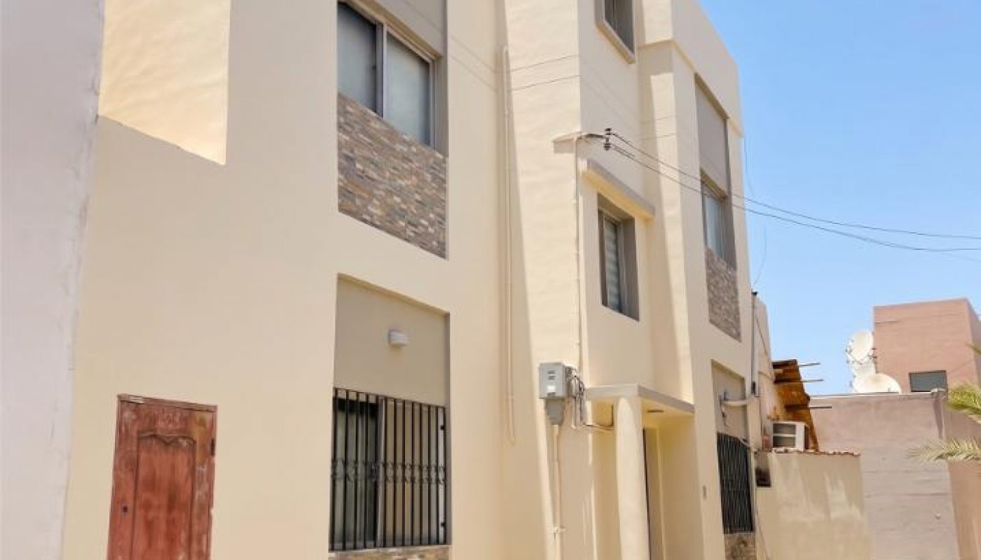 A two-story beige building with stone accents, barred windows, and a brown door, situated on a paved area under a clear, blue sky.