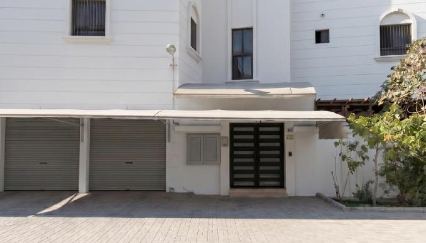 A modern white three-story building with arched windows, two closed garage doors, a main entrance with black glass doors, and a small garden area on the right.