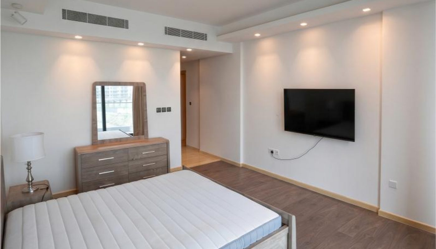 Minimalist bedroom with a wooden bed frame, mattress, dresser with mirror, bedside lamp, wall-mounted TV, and hardwood flooring. Walls and ceiling are white with recessed lighting.