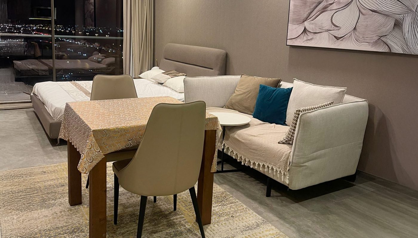 Modern living and dining area with a beige sofa, dining table with two chairs, a patterned rug, wall art, and large window overlooking city lights at night.