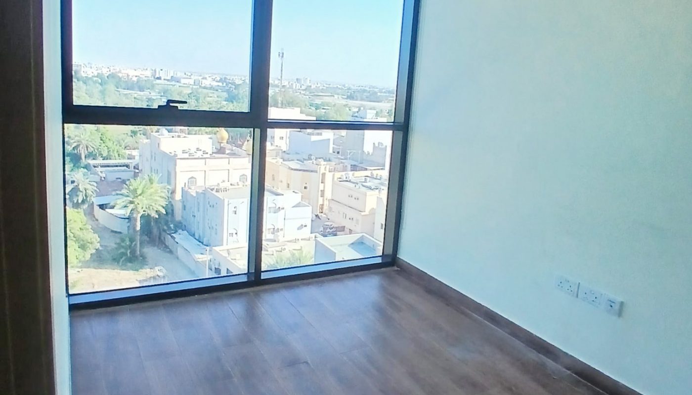 Empty room with wooden floor, large floor-to-ceiling window, and a view of residential buildings and greenery outside in daylight.