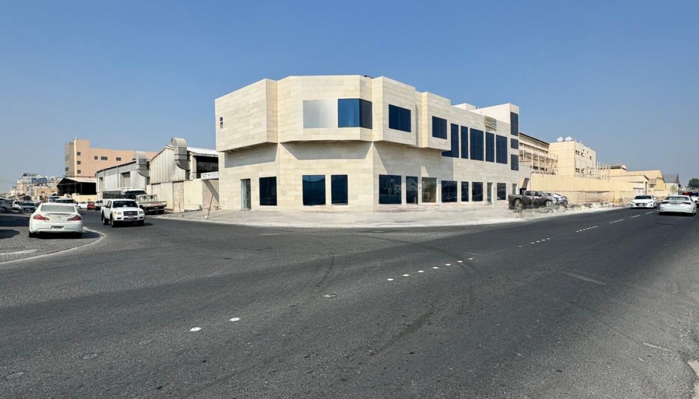 A two-story beige building with large windows on a wide, empty street under a clear blue sky, with several parked cars nearby.