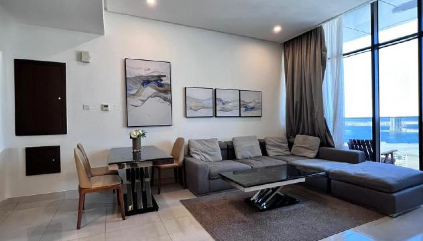 Modern living room with a gray sectional sofa, a small dining area, wall art, and large floor-to-ceiling windows letting in natural light.