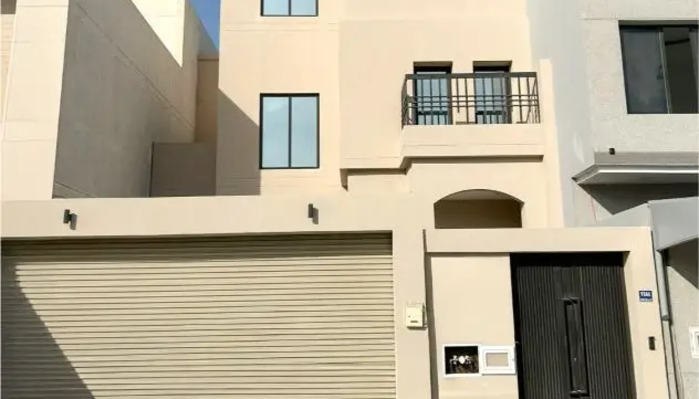 Modern three-story beige house with a closed garage door, black entry gate, and tiled driveway under a partly cloudy sky.