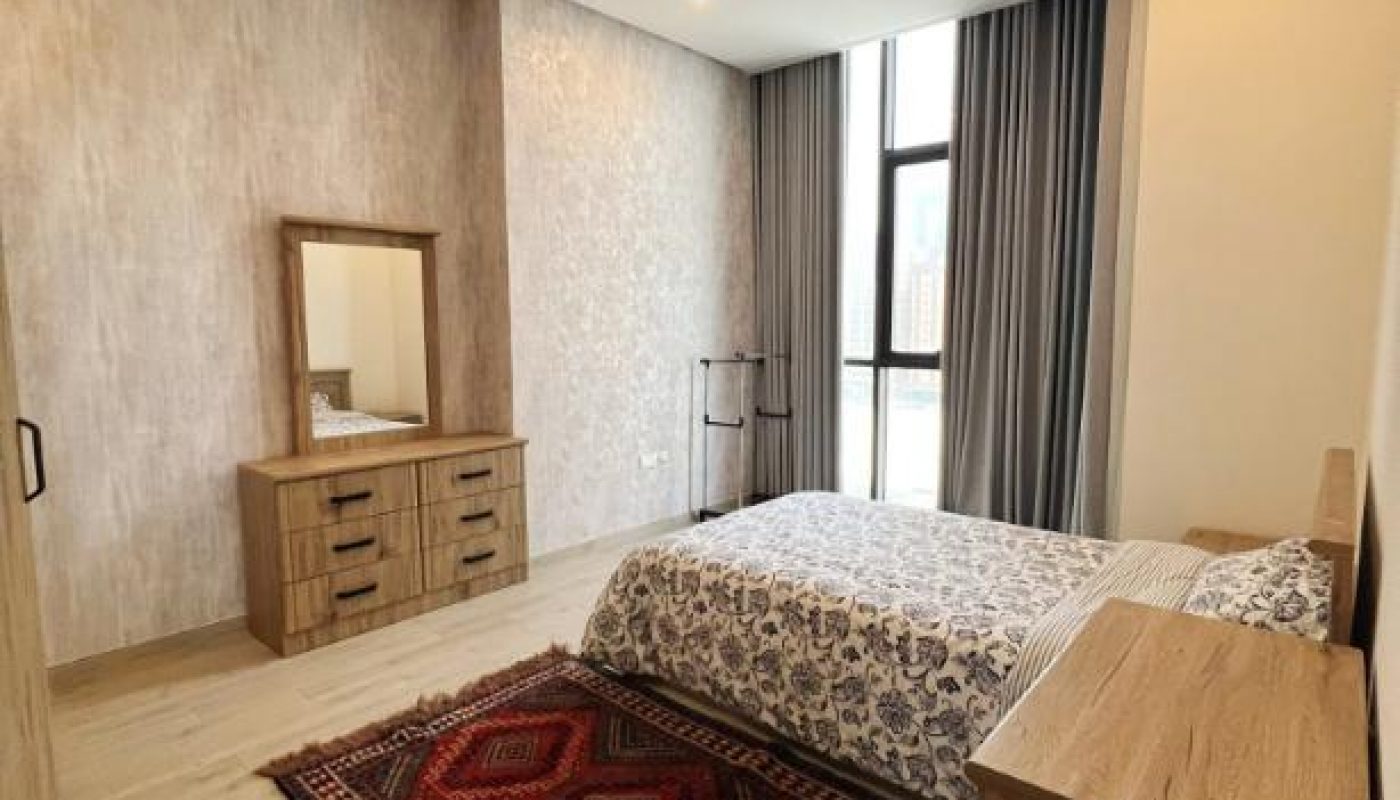 A bedroom with a wooden bed, matching dresser and nightstand, a mirror, patterned bedding, a red rug, and large window with curtains.