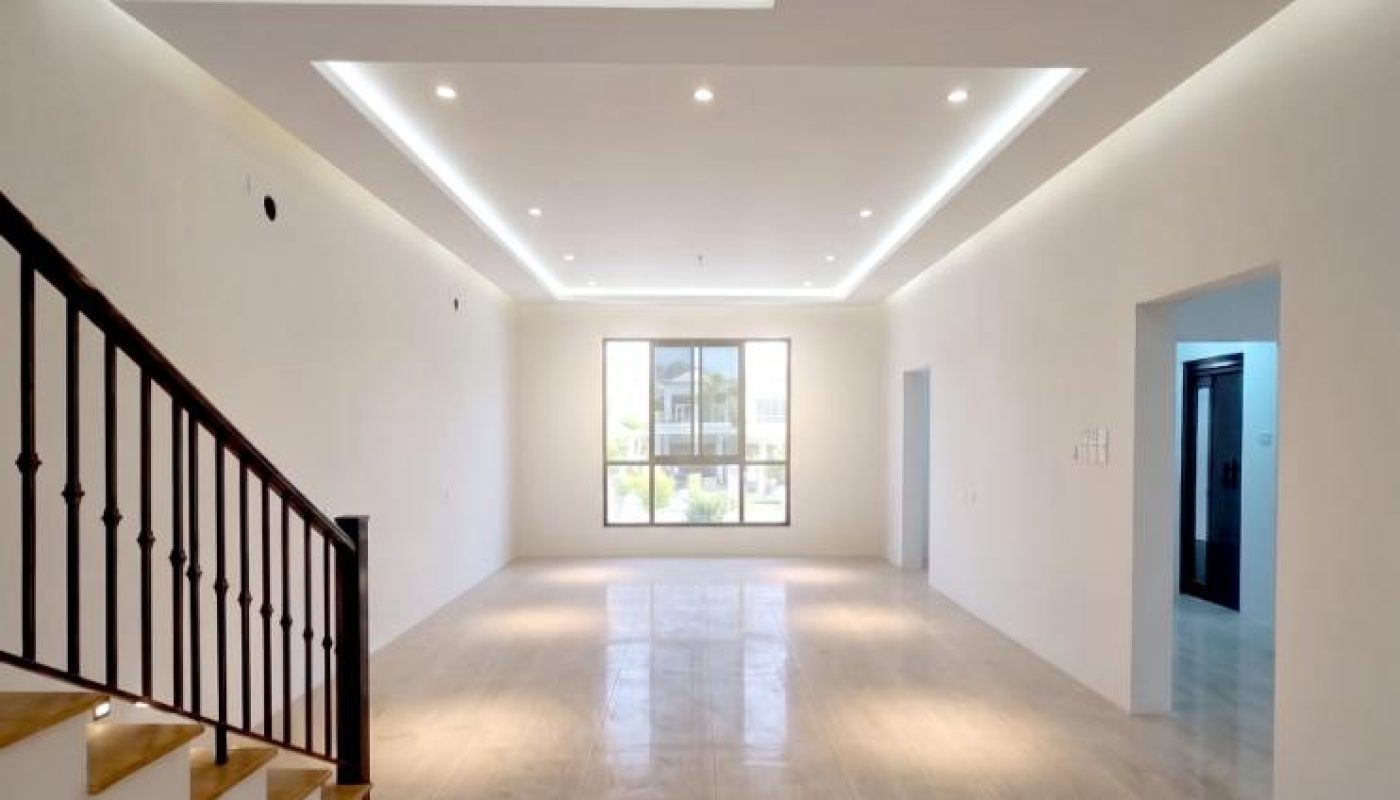 Empty, modern living room with marble floors, recessed ceiling lights, large window at the far end, and a wooden staircase on the left.
