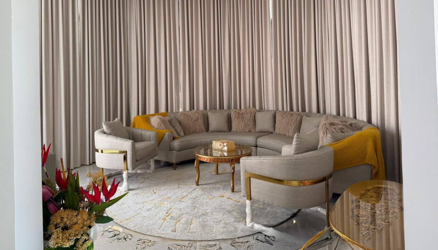 Elegant living room with a large beige sectional sofa, round coffee table, chandelier, gold accents, floor-to-ceiling curtains, and a flower arrangement on the left.