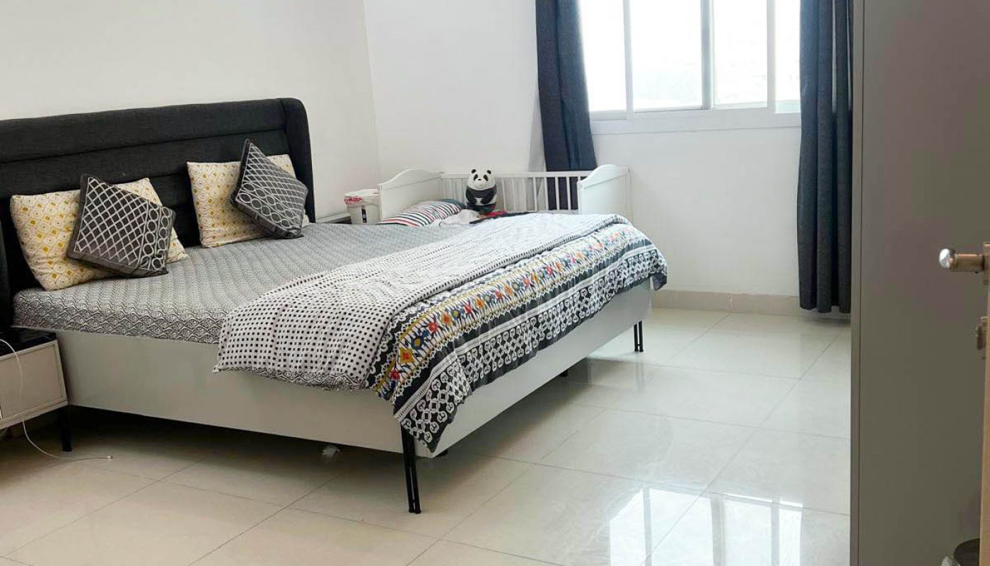 A neatly arranged bedroom with a double bed, gray headboard, patterned bedding, a clock on the wall, a window with dark curtains, and a ceiling fan.