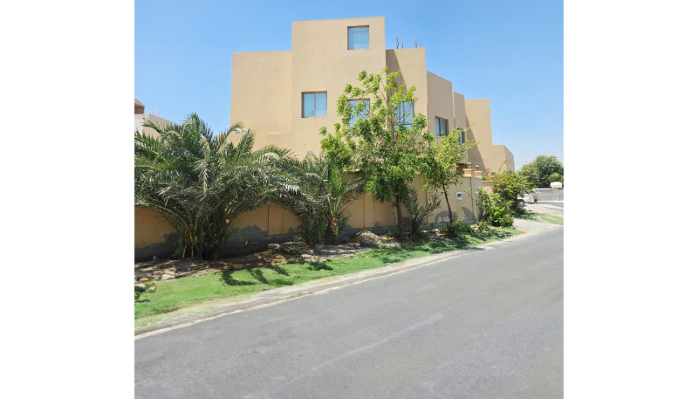 Modern beige building with rectangular windows, surrounded by palm trees and greenery, situated beside a paved road under a clear blue sky.