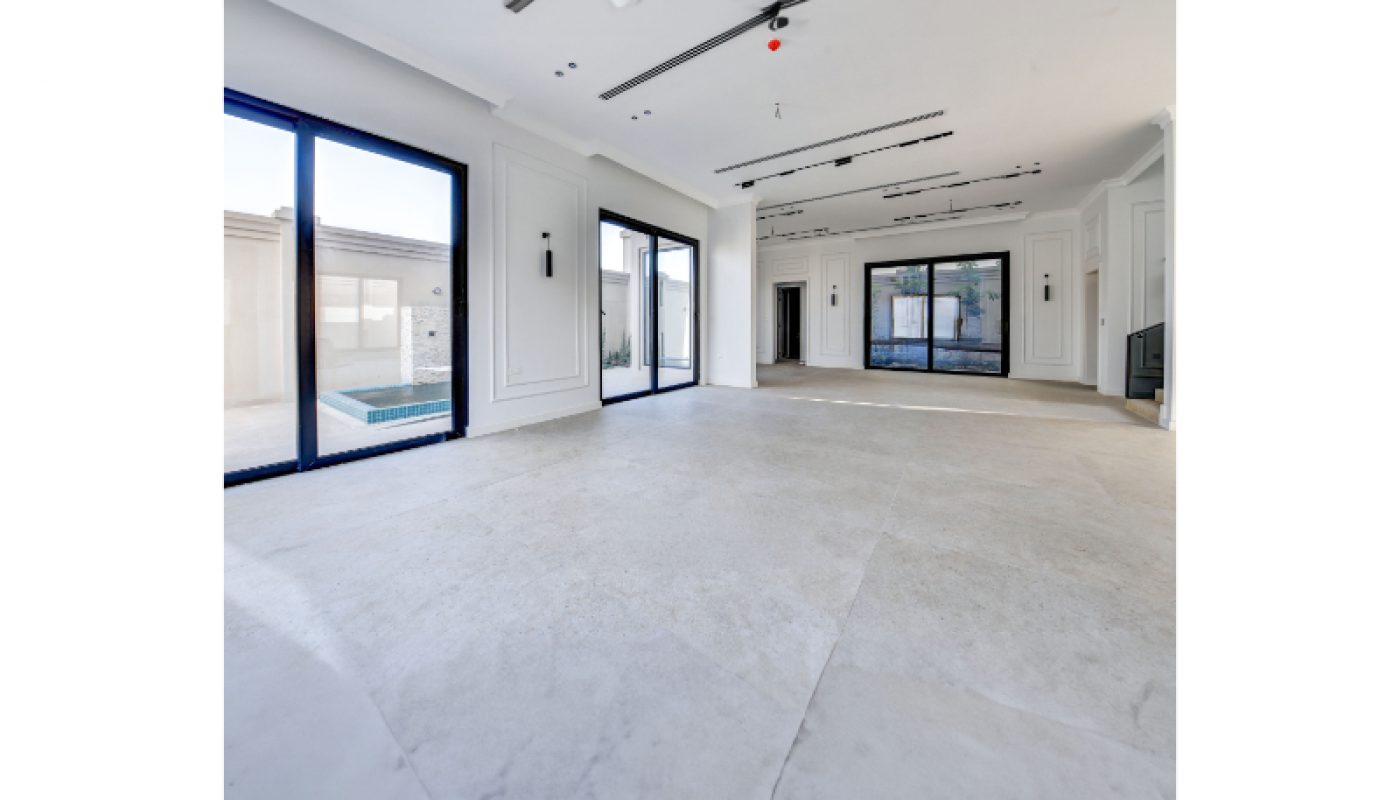 A spacious, empty modern room with large floor-to-ceiling windows, white walls, marble flooring, and views of an outdoor area with a pool.