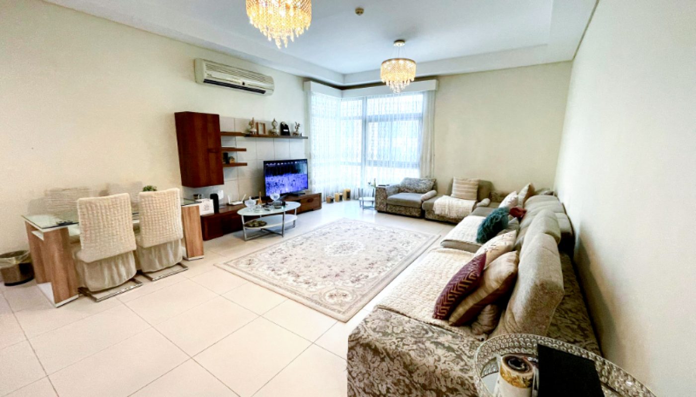 A spacious, well-lit living room with a large L-shaped sofa, TV, dining area, chandeliers, and floor-to-ceiling windows with sheer curtains.