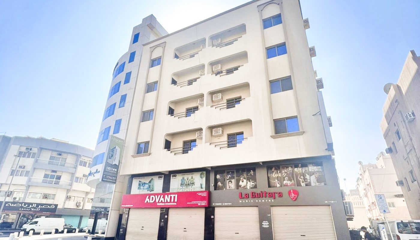A mid-rise beige building on a city street with ground-floor shops, including Advanti and La Bullarys, under a clear blue sky.