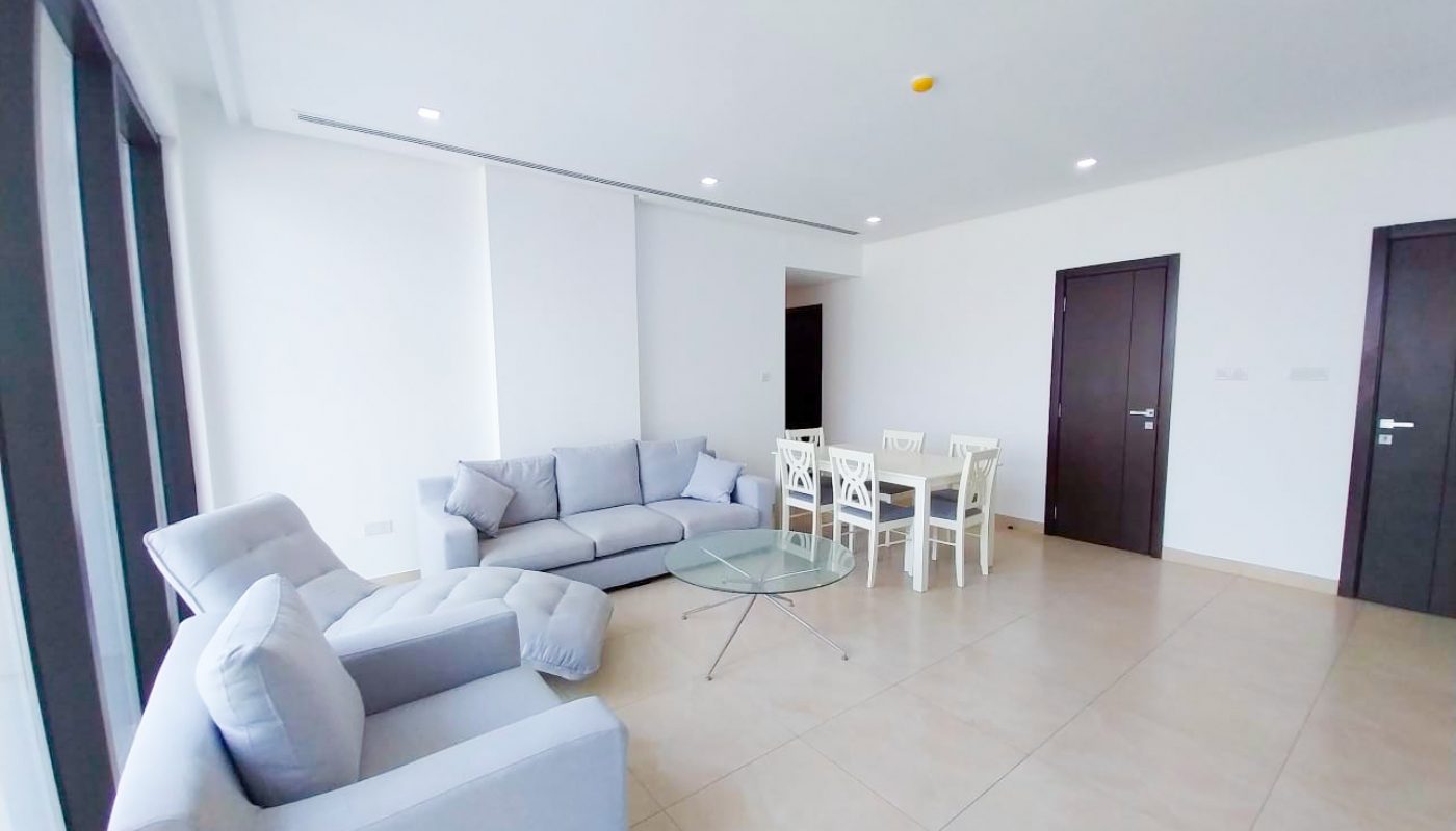 A spacious living and dining area with light-colored sofas, a glass coffee table, a white dining set, tiled floors, and dark-framed doors.