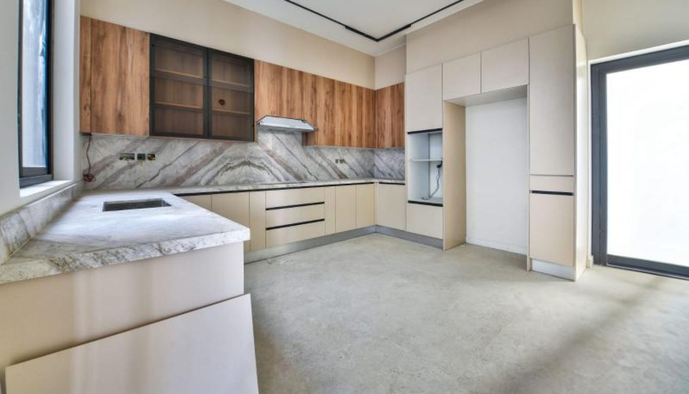Modern kitchen with beige cabinets, marble countertops, wood accents, built-in appliances, and large windows providing natural light. The space is empty and unfurnished.