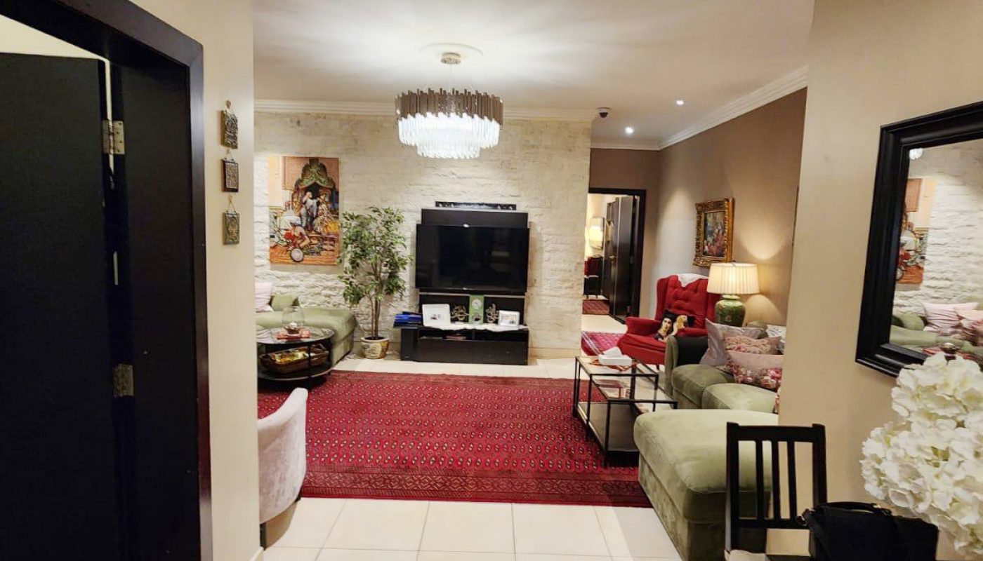 A well-lit living room with sofas, a TV, art on the walls, plants, and red patterned rugs on a tiled floor.