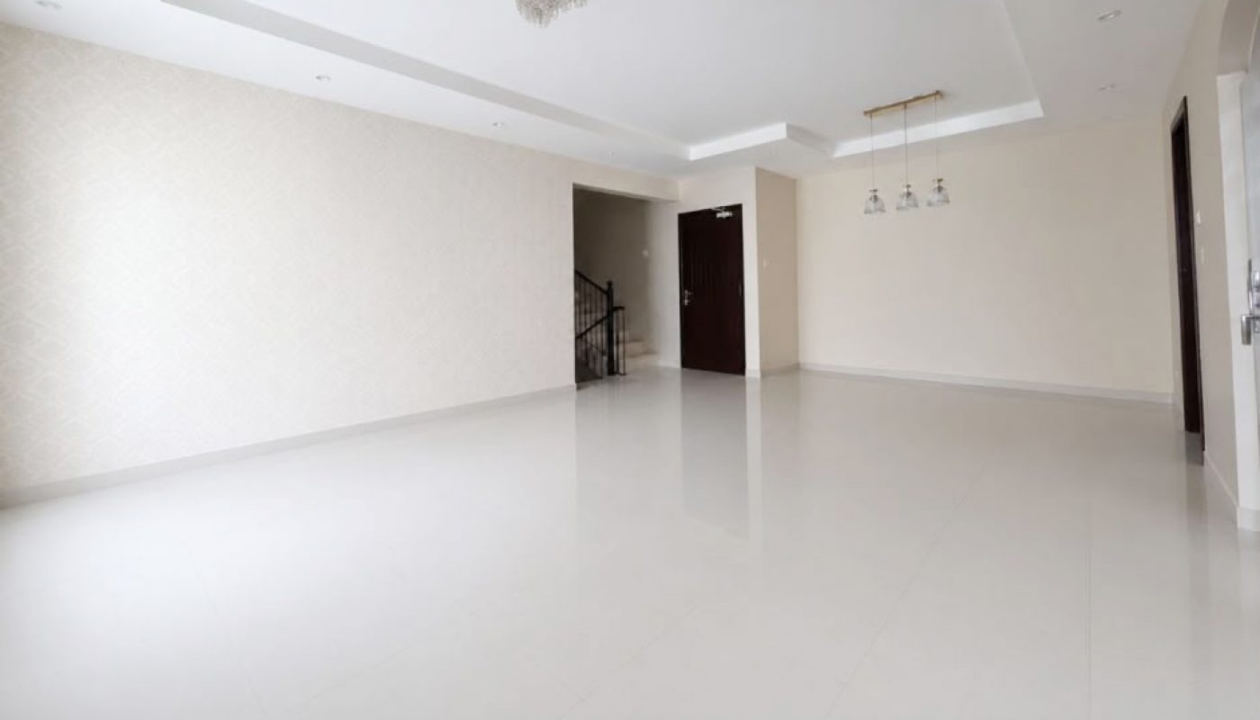 Spacious empty room with white tiled floors, off-white walls, ceiling lights, and a staircase visible near a dark wooden door.