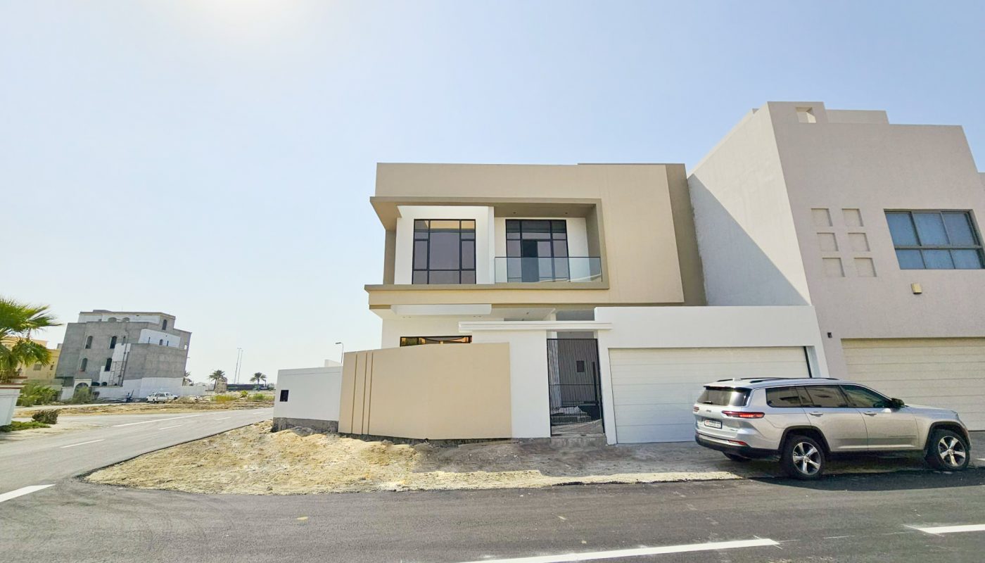 A modern two-story house with a balcony and attached garage, a silver SUV parked in the driveway, and a neighboring unfinished building visible.