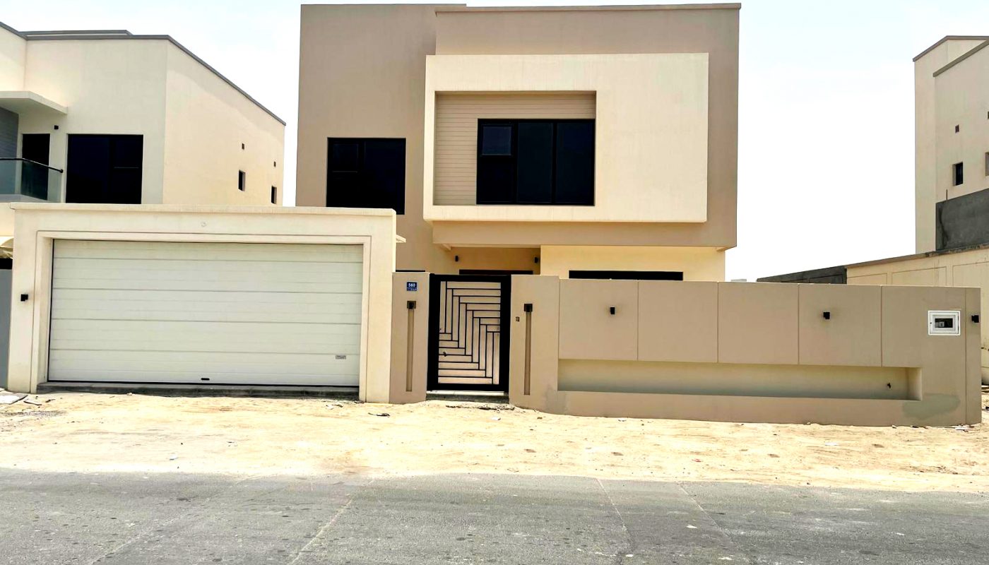 A modern two-story house with a flat roof, large garage door, central entrance gate, and minimalistic beige exterior, located on a sandy plot.