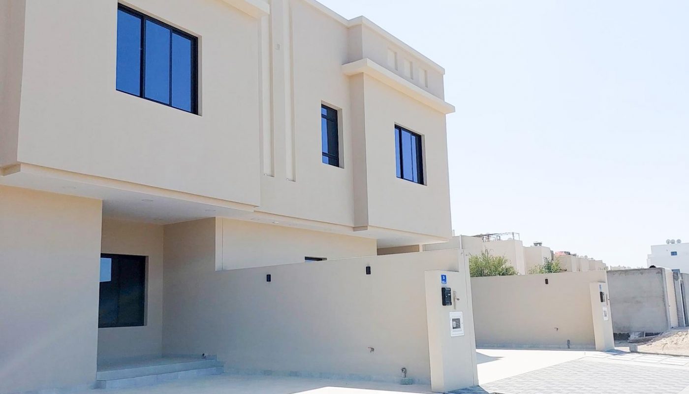 A modern beige two-story house with large windows, a paved driveway, and minimal landscaping under a clear sky.