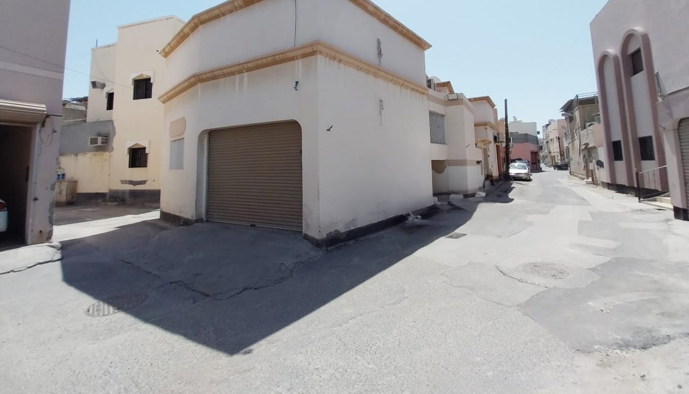 A sunlit, empty street with a two-story white building featuring a closed garage door at the corner. Other buildings line both sides of the street under a clear sky.