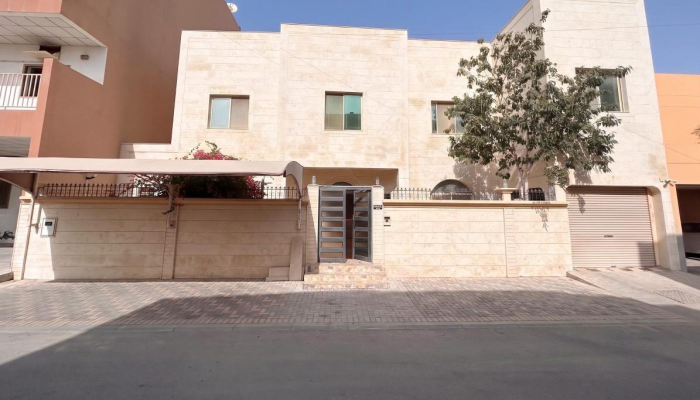A modern two-story beige house with a flat roof, garage, front gate, small tree, and minimal landscaping, set on a paved street under a clear blue sky.