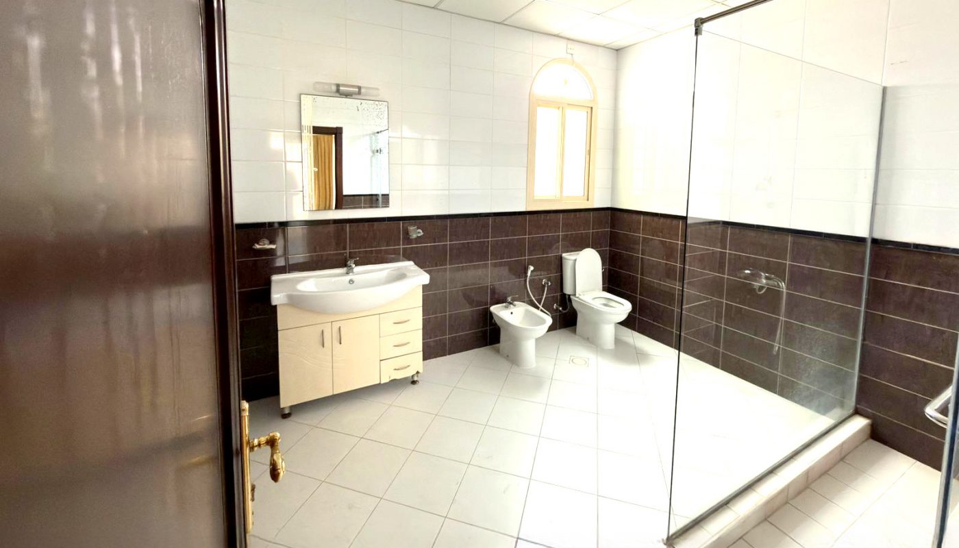 Bathroom with white tile floor, dark lower wall tiles, a sink with vanity, a bidet, a toilet, and a glass-enclosed shower area. A window lets in natural light above the fixtures.