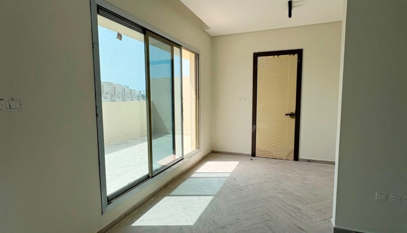 Empty room with light-colored walls, large sliding glass doors to a patio, wood-patterned tile flooring, and a door covered in protective plastic. Sunlight is streaming in through the glass doors.