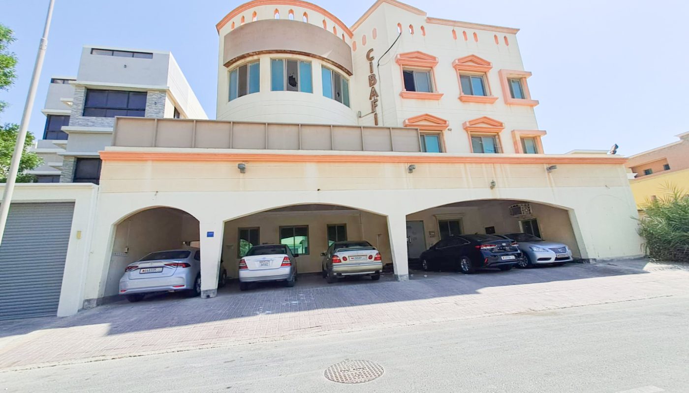 A three-story beige building with orange trim, labeled "Gibal," featuring arched parking spaces occupied by cars, seen from the street on a sunny day.