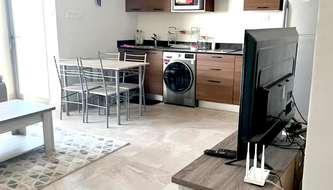 Modern open-plan kitchen and living area with brown cabinets, a washing machine, dining table, fridge, and a TV on a light wood stand. A clock hangs above the kitchen.