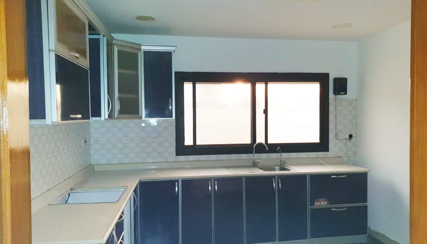 An empty kitchen with blue cabinets and a window in a cozy apartment for sale in Busaiteen.