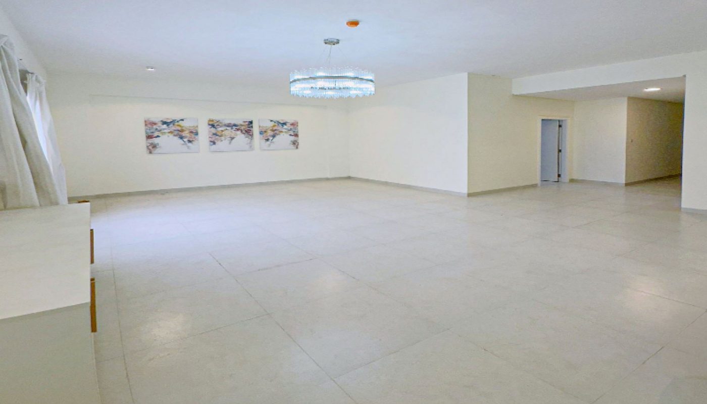 Spacious empty room with white tiled floor, three abstract paintings on the wall, a chandelier, large window with curtains, and a door in the far right corner.