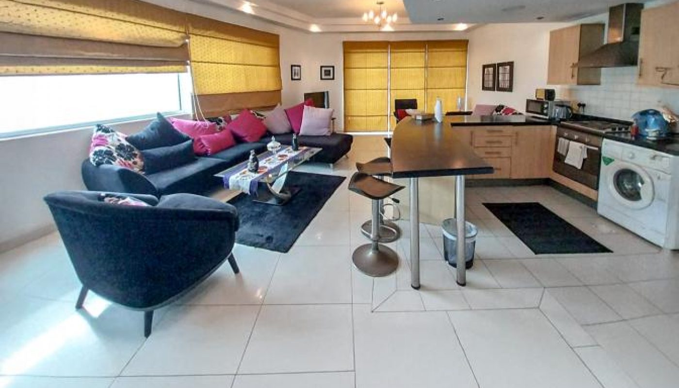 Modern open-plan living area with a corner sofa, kitchen, island with bar stools, and large windows with yellow blinds. Laundry machine and cabinets visible.