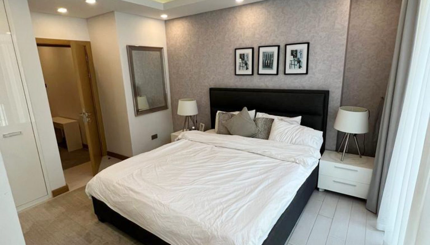 Modern bedroom with a neatly made bed, two bedside tables with lamps, three black-and-white photos above the headboard, and a large window with sheer curtains.